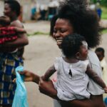 A smiling mother holding her baby in one arm and a meal in the other, symbolizing hope and support provided by Hunger Relief West Africa.